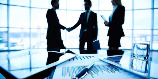 Two business people shaking hands whilst one looks on. In the forefront are some papers on a desk.